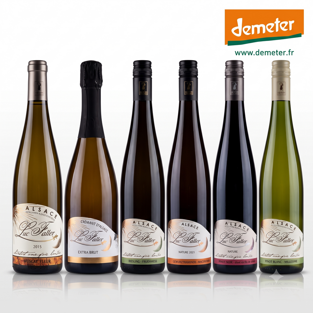 Six bottles of Alsace Vignoble Luc Faller  wines r, arranged in a row against a white background. The bottles have different colored glass (green, gold, dark brown, black) and different colored labels, with the Demeter logo visible in the top right corner.