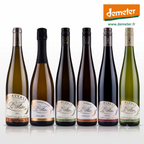 Six bottles of Alsace Vignoble Luc Faller  wines r, arranged in a row against a white background. The bottles have different colored glass (green, gold, dark brown, black) and different colored labels, with the Demeter logo visible in the top right corner.