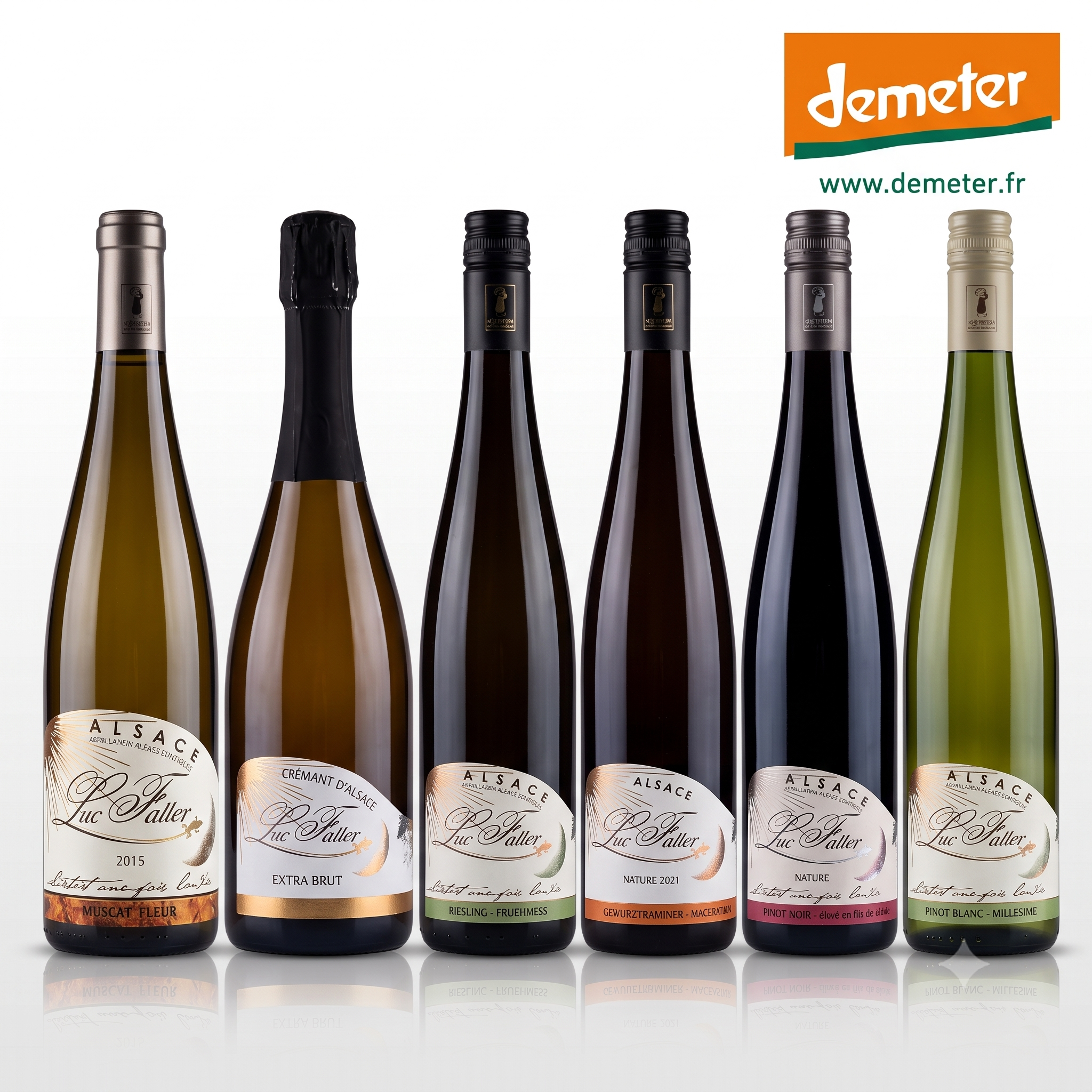 Six bottles of Alsace Vignoble Luc Faller  wines r, arranged in a row against a white background. The bottles have different colored glass (green, gold, dark brown, black) and different colored labels, with the Demeter logo visible in the top right corner.