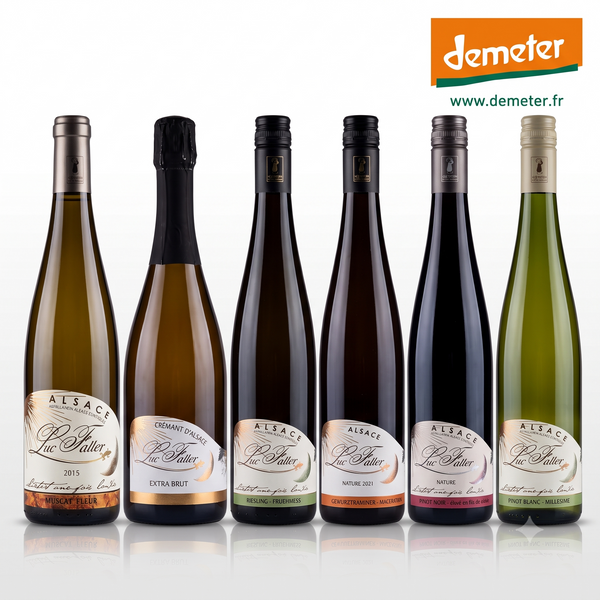 Six bottles of Alsace Vignoble Luc Faller  wines r, arranged in a row against a white background. The bottles have different colored glass (green, gold, dark brown, black) and different colored labels, with the Demeter logo visible in the top right corner.