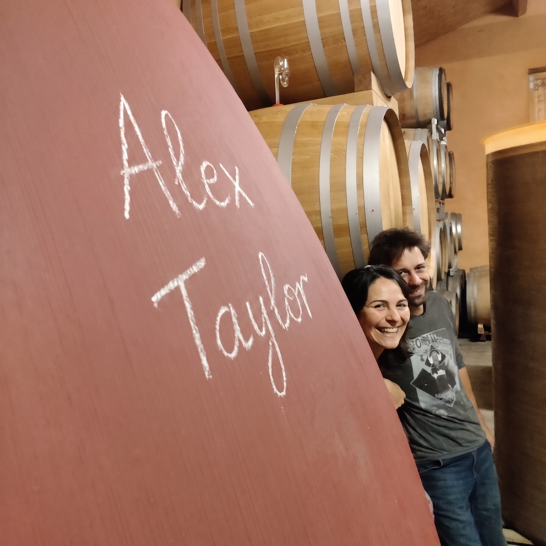 Flo and Paola  standing behind a large wooden barrel with 'Alex Taylor' written on it, surrounded by wine barrels.