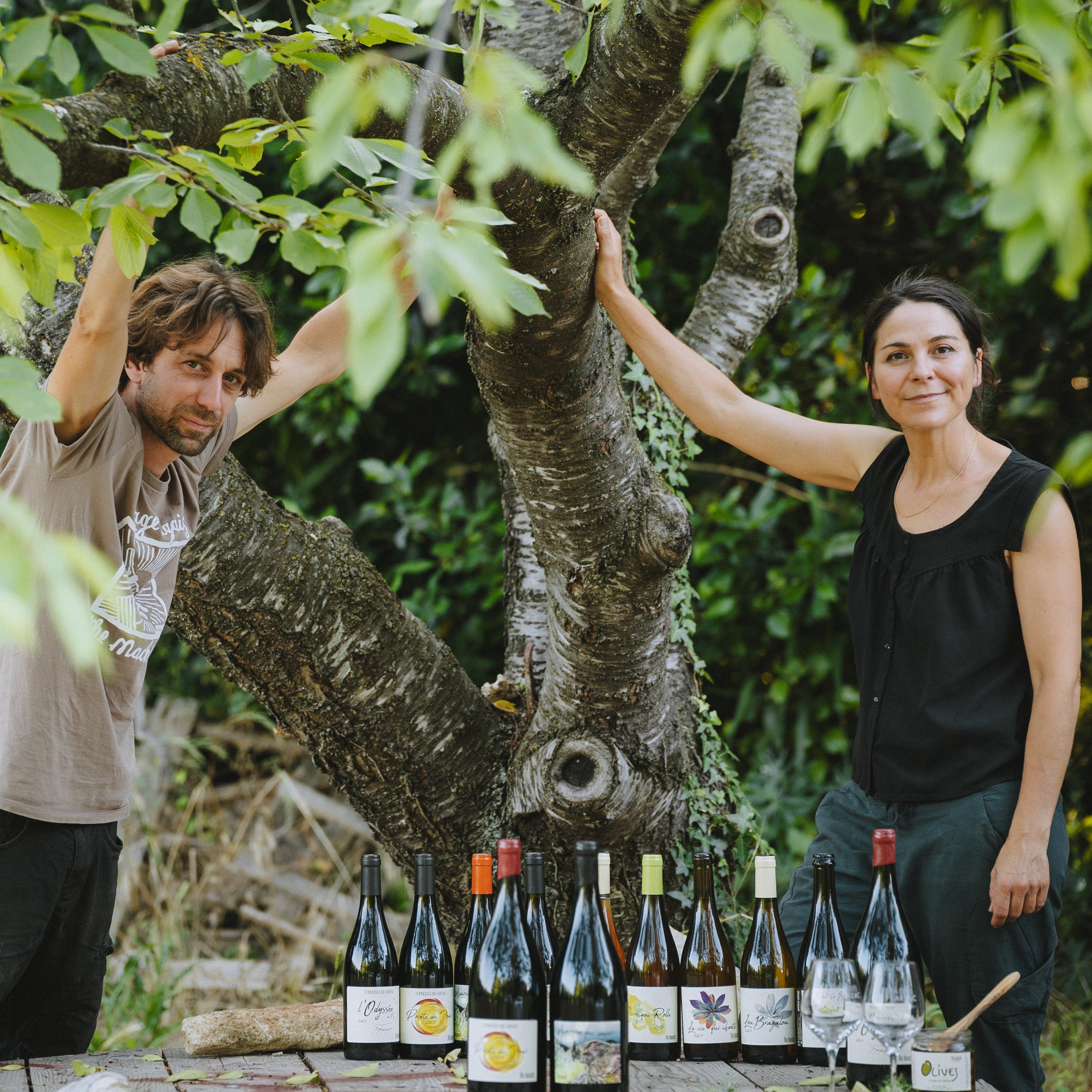 Paola and Flo standing outdoors with a table of wine bottles under a tree at DomainecFloBusch