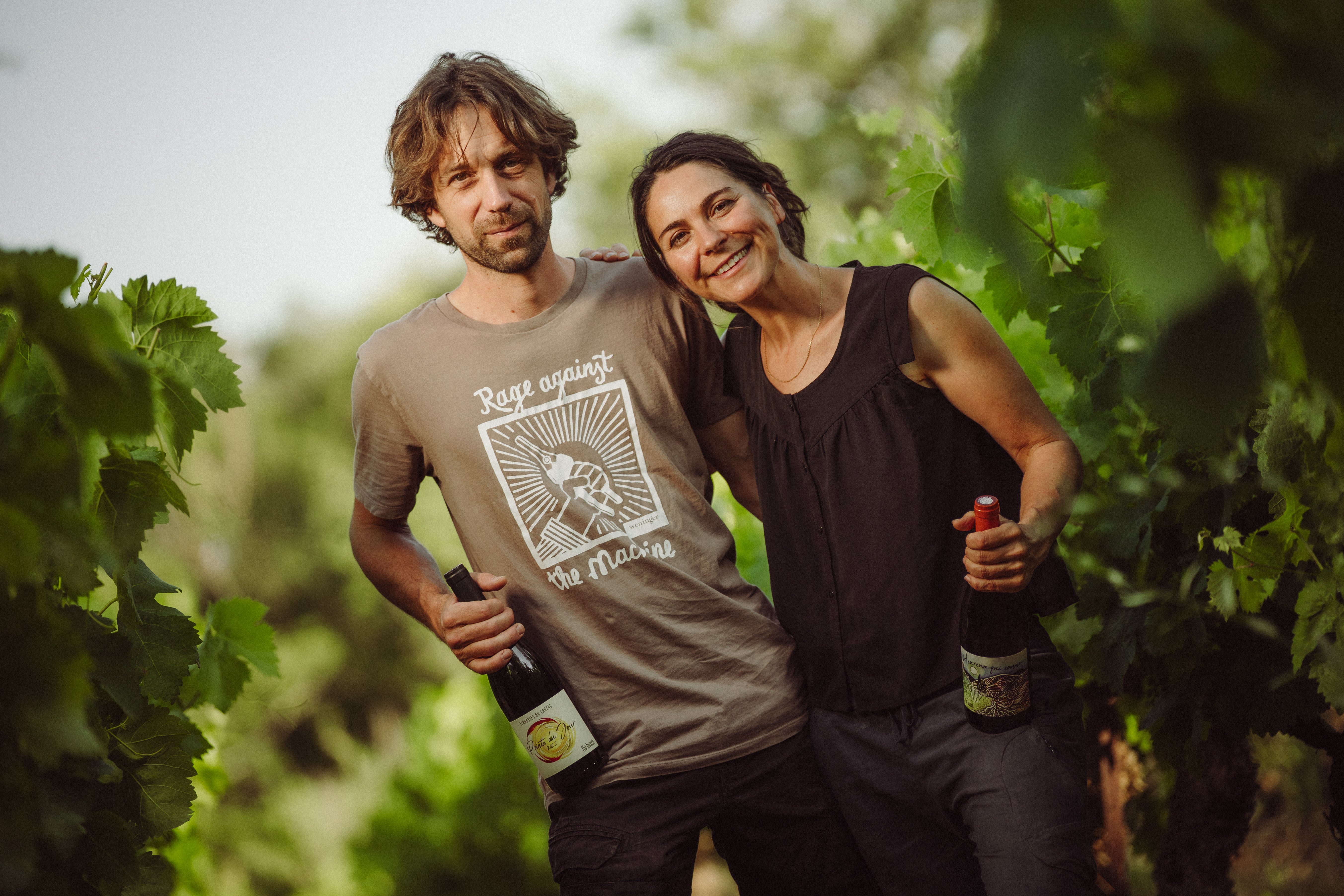 Two people standing in a vineyard holding wine bottles.