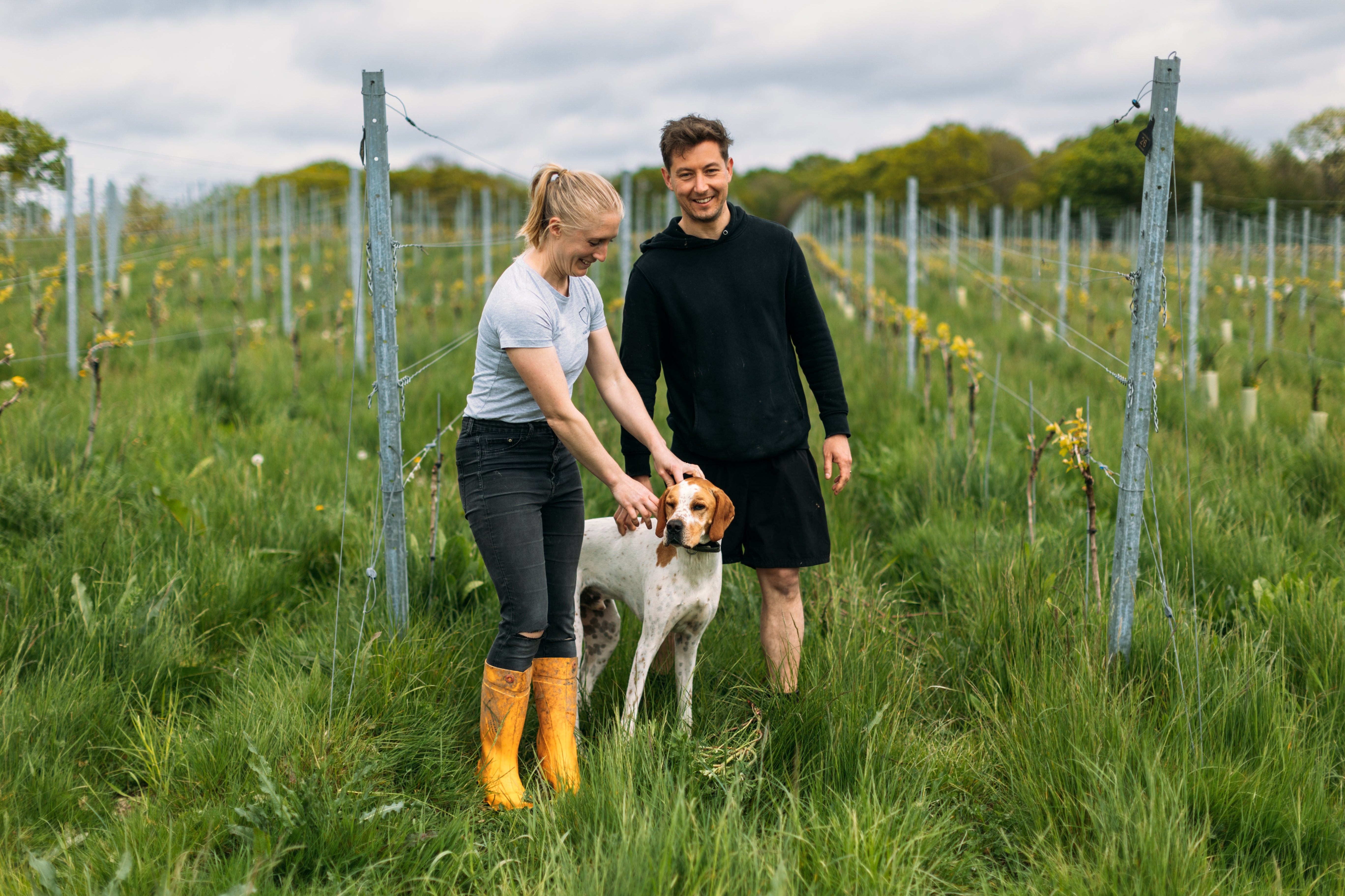 A man and a woman standing in a vineyard with a white and brown dog, wearing yellow rain boots, a light blue t-shirt, and a black long-sleeve top respectively.