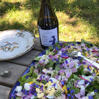 An outdoor dining scene on a wooden table with a large blue platter filled with a colorful salad of greens, purple and white flowers, and sliced radishes. A dark wine bottle with a white label reading 'Pure Flower Wine' stands behind the platter, alongside plates and a spoon, with grass in the background.