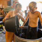 A woman and a young boy participate in a wine-making process, with the woman pouring dark grapes from a black bucket into a large hammered metal vat while the boy stands inside the vat.