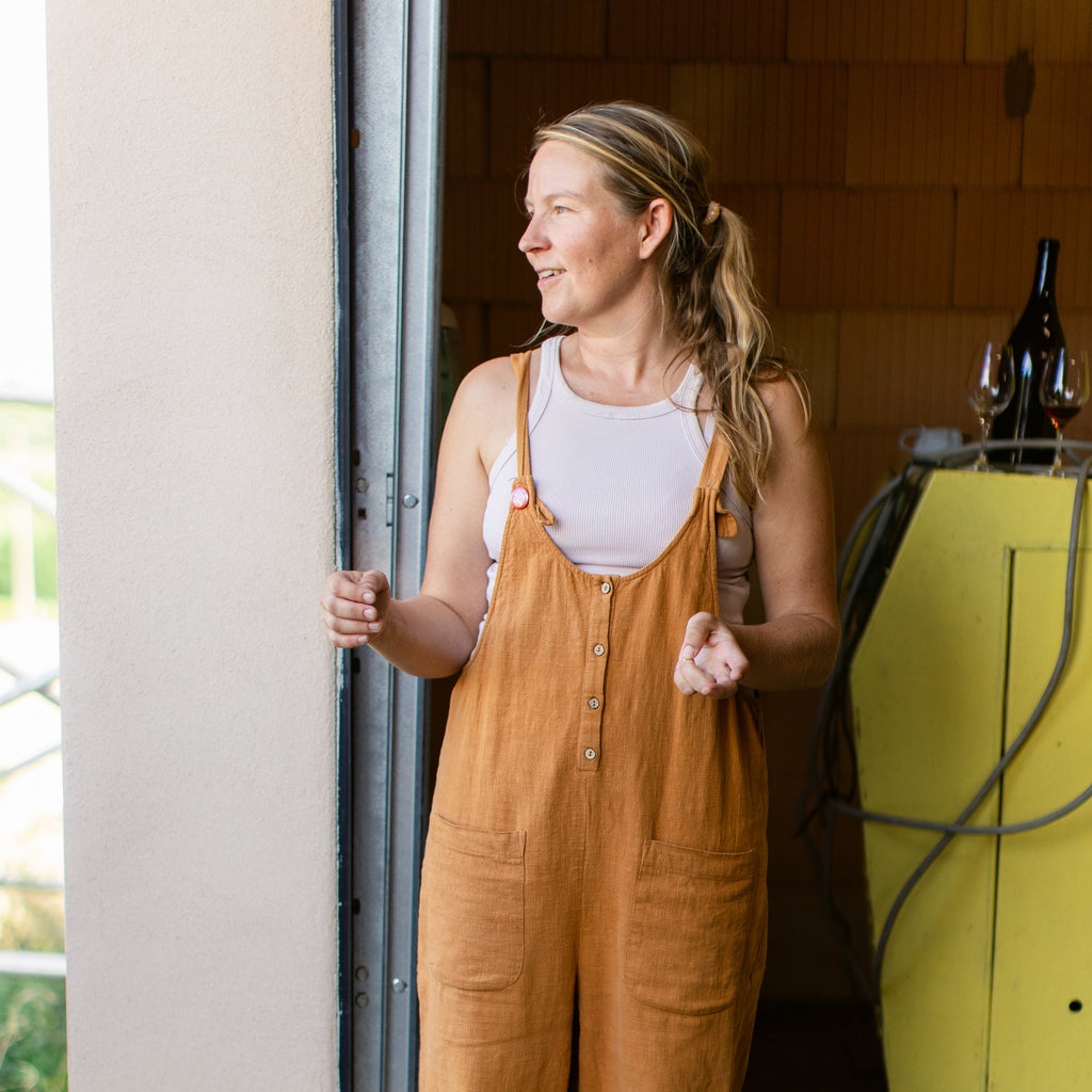 A woman wearing a pair of wide-leg mustard brown linen utility overalls over a white tank top, standing in a garage doorway.