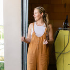 A woman wearing a pair of wide-leg mustard brown linen utility overalls over a white tank top, standing in a garage doorway.