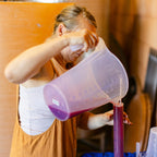 A woman in a workshop pours a purple liquid from a translucent plastic pitcher into a tall glass container on a blue cloth-covered table.