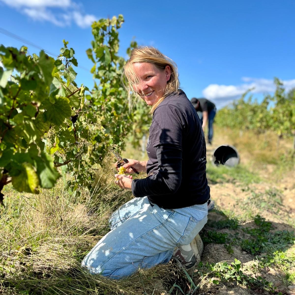 A Anne Lekkerkerker kneeling in a vineyard under a bright blue sky, smiling at the camera while holding a small object. She is wearing a dark long-sleeve top and light blue jeans, with grapevines and another person visible in the background.