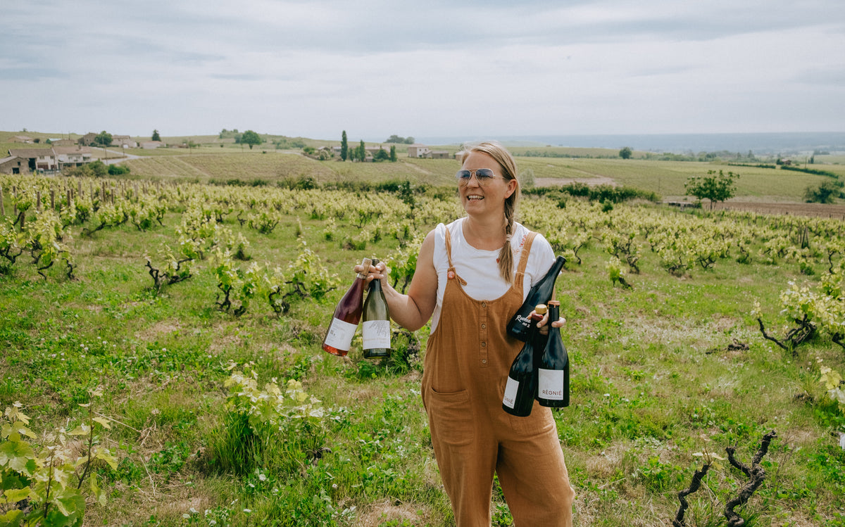 A woman stands in a vineyard holding several wine bottles, wearing brown overalls over a white top, with rows of grapevines and a cloudy sky in the background.