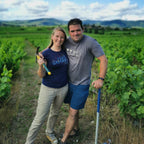 A man and a woman standing in a vineyard field under a blue sky with clouds. The woman wears a navy blue t-shirt with 'JAKE'S SAMHAY' printed on the front, paired with beige pants, while the man wears a gray t-shirt and blue shorts.