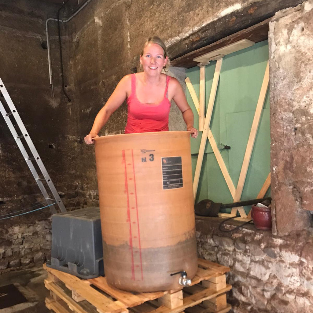 A woman in a pink tank top stands behind a large wooden barrel-style container placed on wooden pallets in a rustic stone room with a green door in the background.