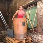 A woman in a pink tank top stands behind a large wooden barrel-style container placed on wooden pallets in a rustic stone room with a green door in the background.