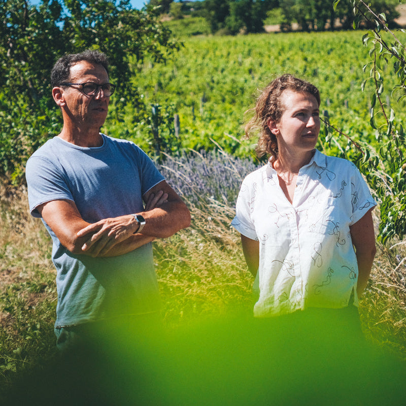 Nicolas in a blue t-shirt and a Marion Lebrun in a white button-up shirt standing in a vineyard field, viewed through a blurry green foreground leaf.