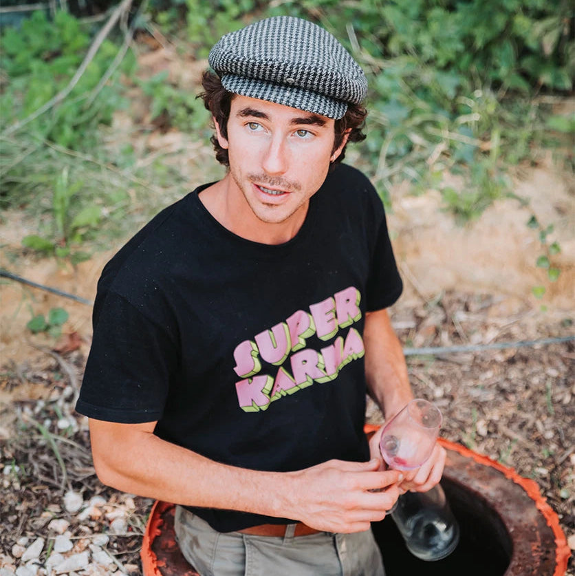 Raf Raybois, wearing a black t-shirt with 'Super Karma' text, sitting on a qvevri  outdoors.