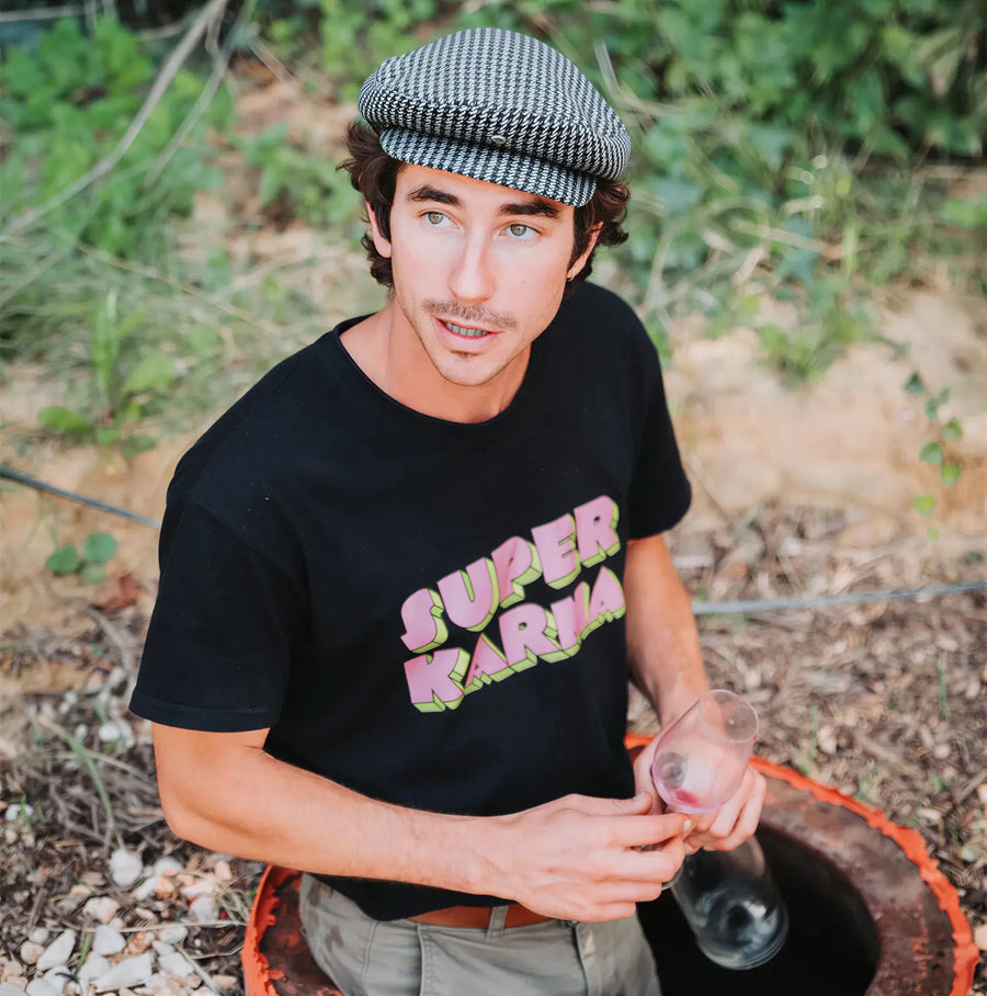 Raf Raybois, wearing a black t-shirt with 'Super Karma' text, sitting on a qvevri  outdoors.