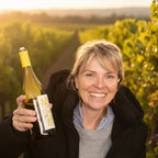 A smiling woman with short blonde hair, Ingrid Nueil holds up a bottle of white wine in a black jacket over a gray sweater against a plain white wall.