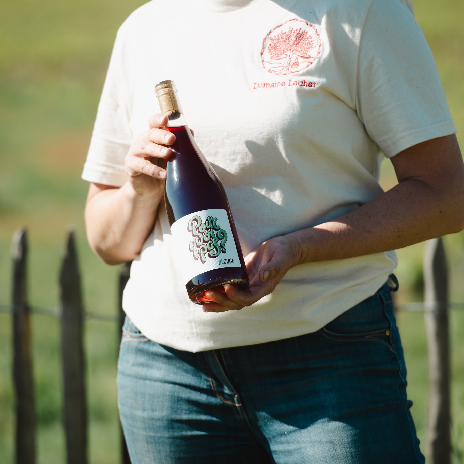 A person outdoors wearing a white t-shirt with a red circular logo and holding a dark wine bottle with a grape label.