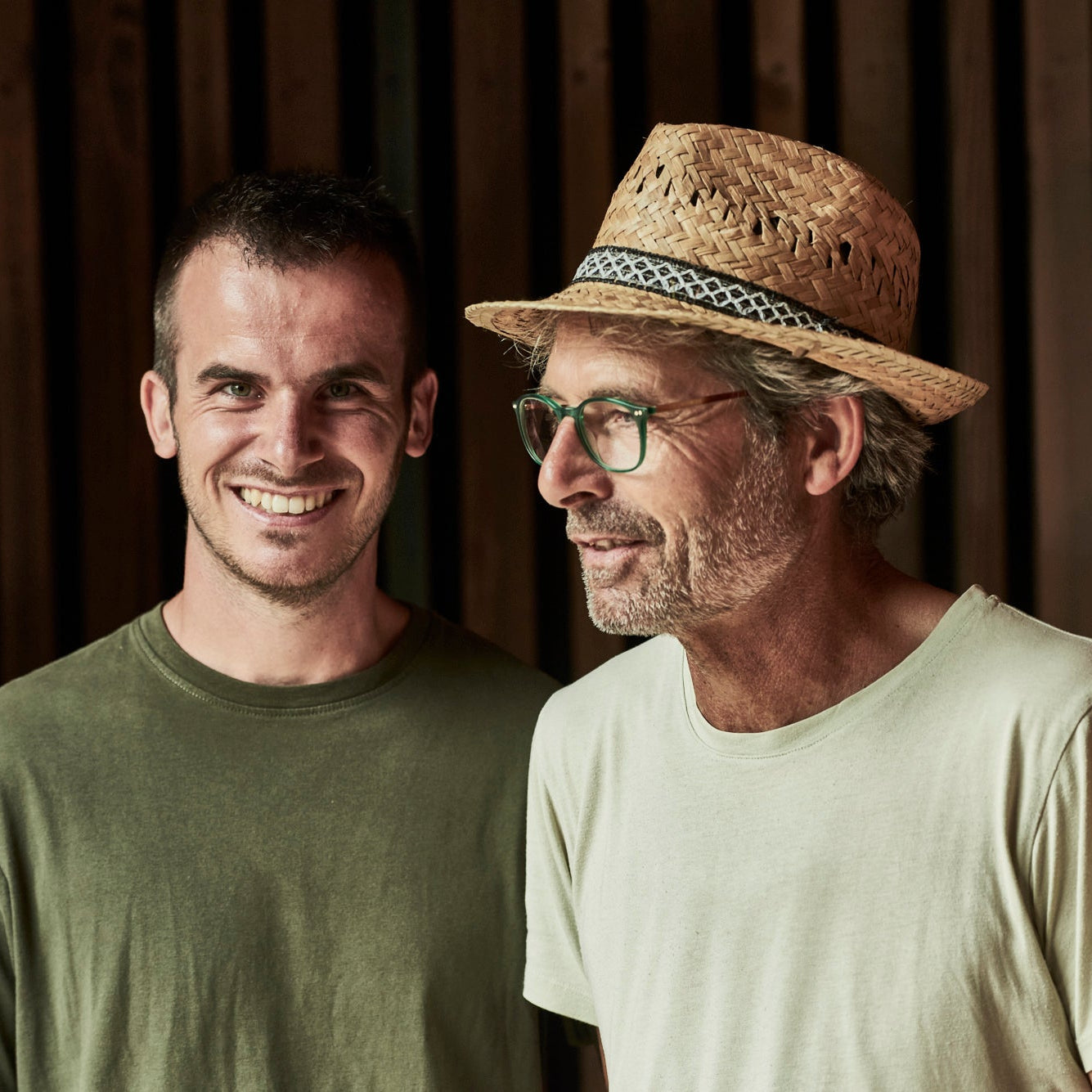 Joachim Unterhofer and Christoph Unterhofer  standing side by side against a dark wood slat wall. The man on the left wears an olive green crewneck t-shirt, and the man on the right wears a light beige t-shirt, a straw hat with a patterned band, and glasses.