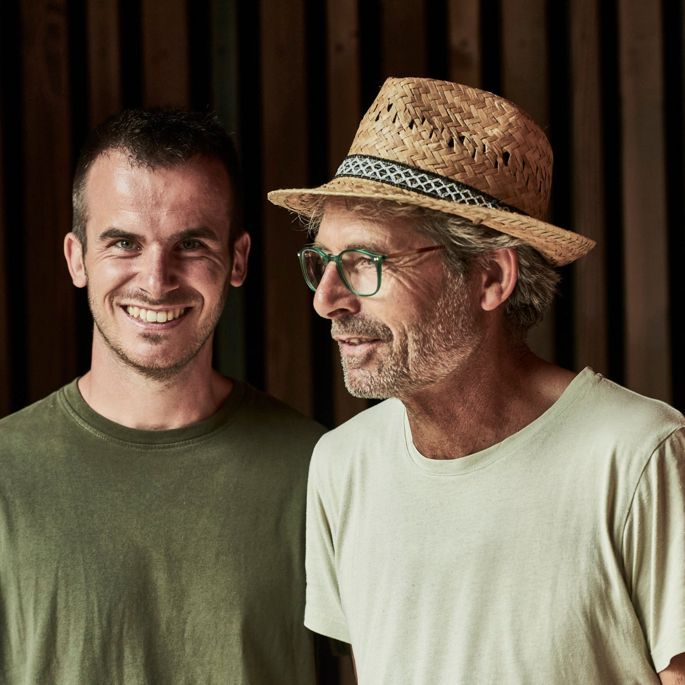 Two men standing side by side against a dark wood slat wall. The man on the left wears an olive green crewneck t-shirt, and the man on the right wears a light beige t-shirt, a straw hat with a patterned band, and glasses.