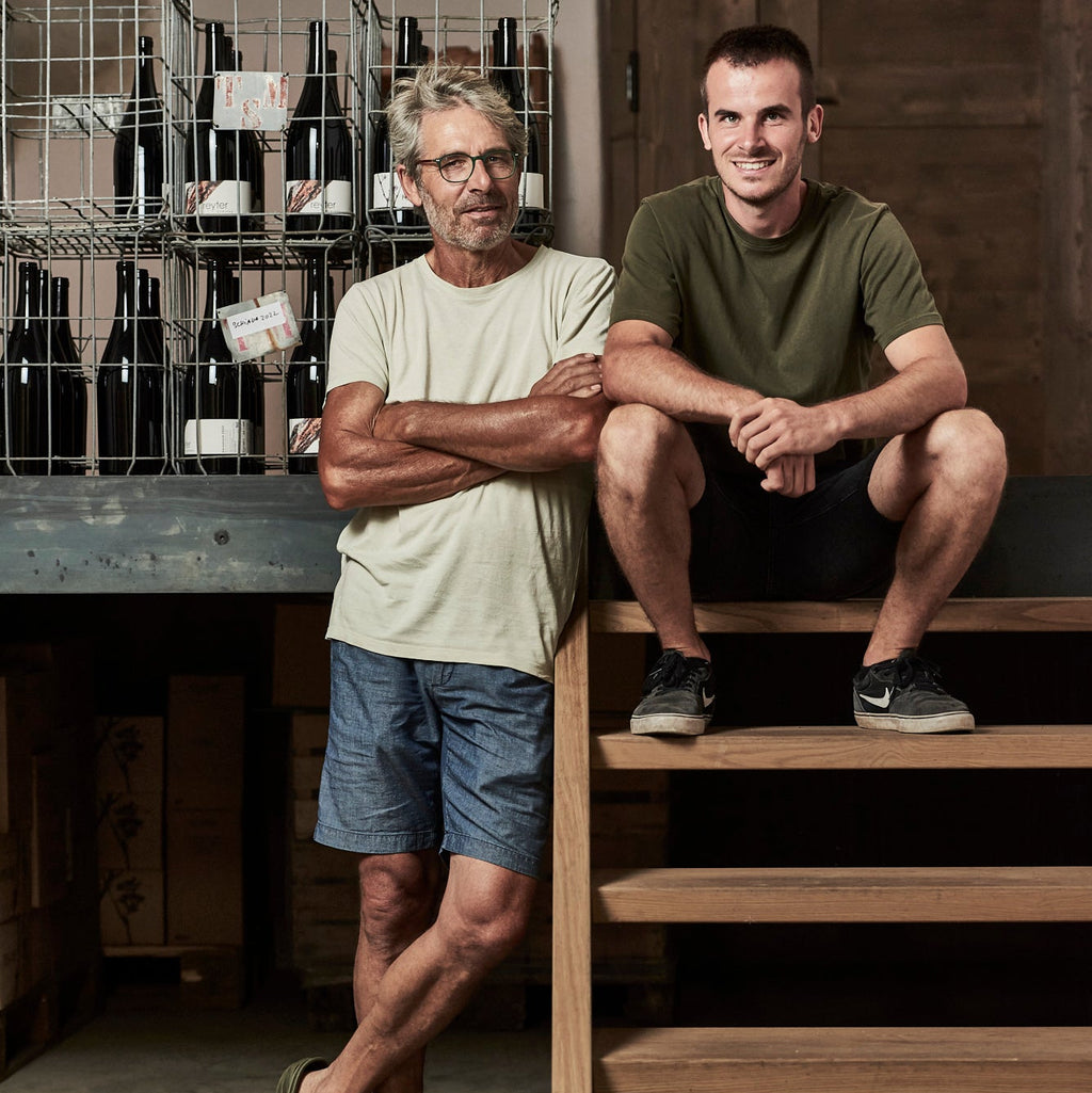 Two men posing in a rustic wine cellar or winery. The man on the left stands wearing a light beige t-shirt and denim shorts, while the man on the right sits on wooden stairs wearing an olive green t-shirt and black shorts.