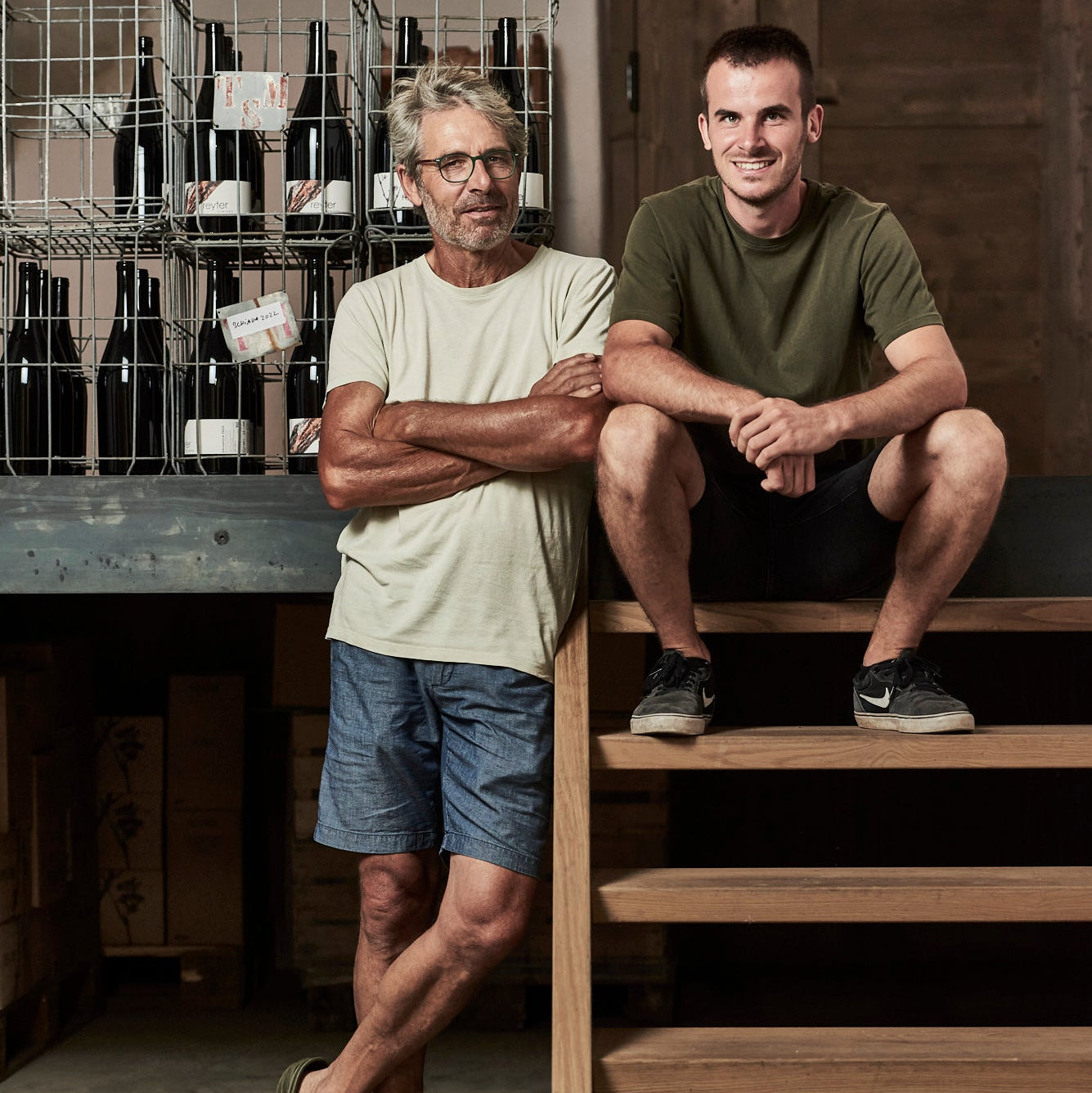 Two men posing in a rustic wine cellar or winery. The man on the left stands wearing a light beige t-shirt and denim shorts, while the man on the right sits on wooden stairs wearing an olive green t-shirt and black shorts.