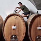 A man in a green t-shirt and black shorts crouches on top of a large wooden wine barrel in a cellar, pouring liquid from a glass container. Another barrel is visible to the right, and a textured stone archway forms the background.