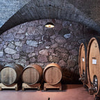 A wine cellar with large wooden barrels lined up against a stone wall under a dark arched ceiling.