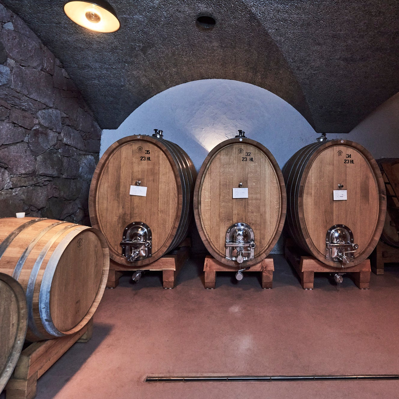 A row of large wooden wine barrels on wooden legs inside a stone-walled cellar with arched ceilings and warm overhead lights.