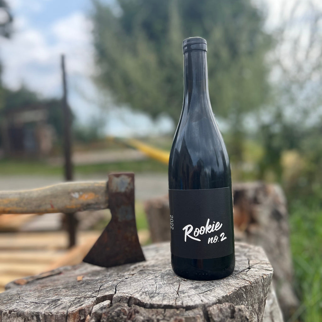 A dark glass wine bottle with a black label reading 'Rookie no. 2' stands on a weathered tree stump outdoors, with a rustic axe beside it and a blurred natural background.