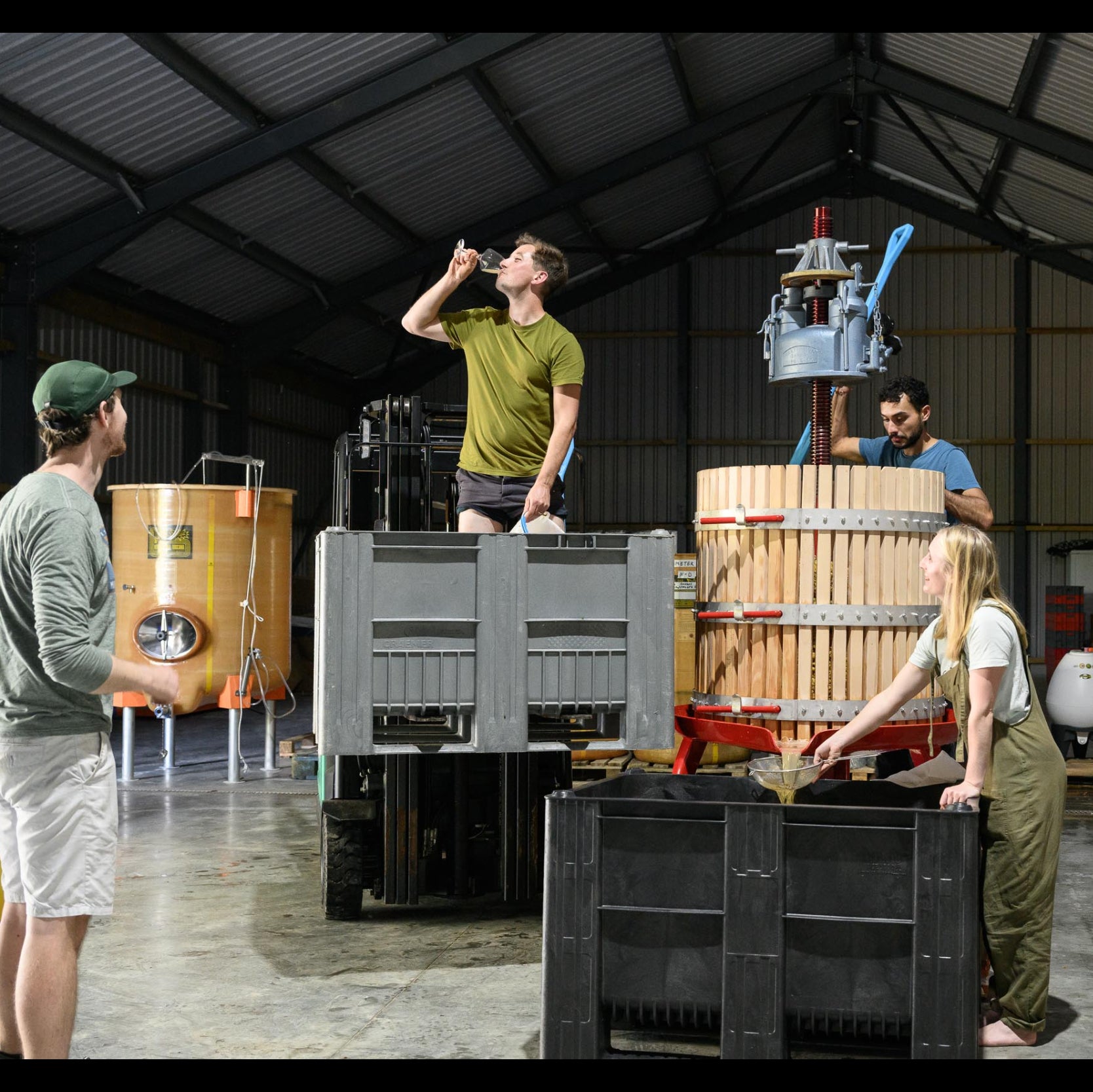 Several people working inside a large industrial warehouse or brewery space, handling a large wooden barrel press with a press machine, black crates, and wooden barrels. A yellow vat is visible in the background.