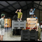 Several people working inside a large industrial warehouse or brewery space, handling a large wooden barrel press with a press machine, black crates, and wooden barrels. A yellow vat is visible in the background.