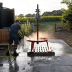 A man uses a hose to rinse a red metal table mounted on a wooden base outdoors. The setup is on a wet concrete patio with a large black water tank, greenery, and a fence in the background.