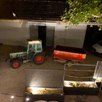 A green tractor with red wheels is parked next to a red trailer in a nighttime outdoor setting, with a garage door, white van, tree branches, and illuminated planter boxes in the foreground.