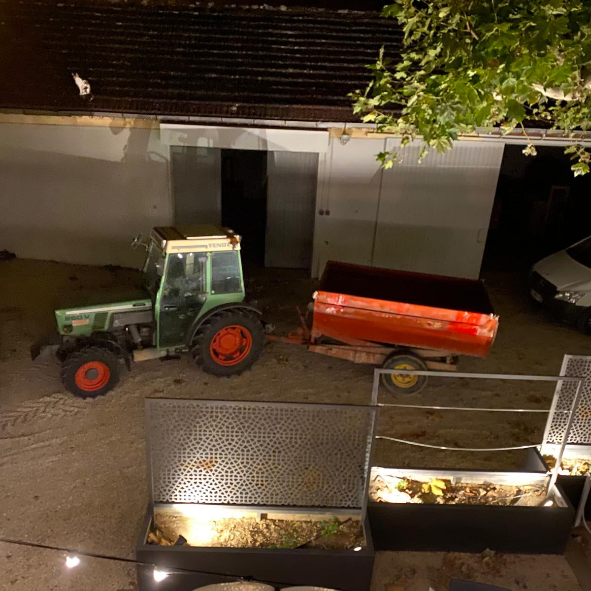 A green tractor with red wheels is parked next to a red trailer in a nighttime outdoor setting, with a garage door, white van, tree branches, and illuminated planter boxes in the foreground.