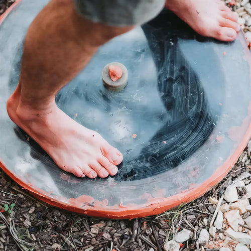 Foot on a round, black qvevri lid set in a gravel area, showcasing the texture and earthy surroundings.